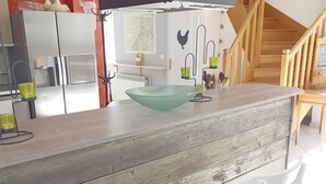A modern kitchen with a central island, white chairs, and a staircase in the background.