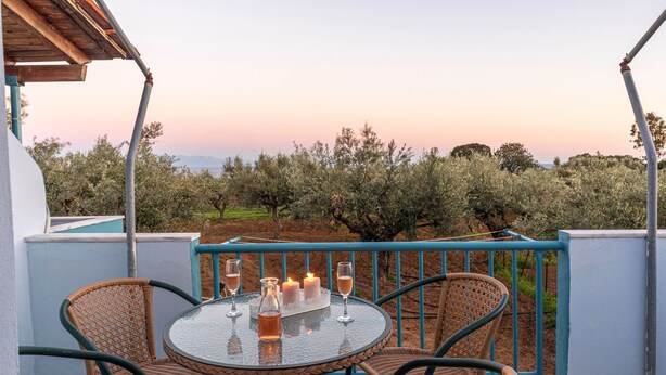 Un tavolo rotondo con sedie in vimini e un piano in vetro, su un balcone affacciato su ulivi e un tramonto.