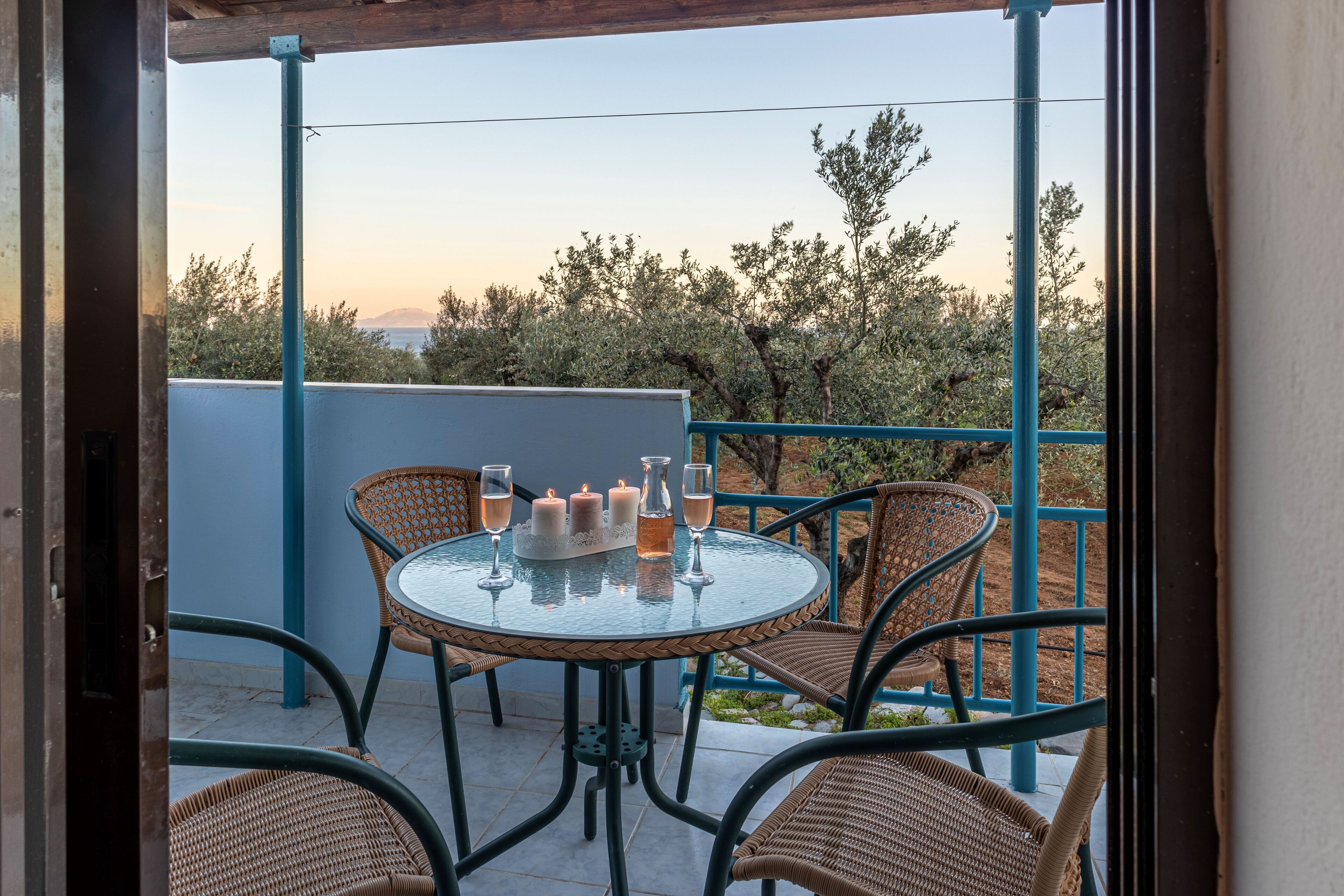 Mesa de vidro redonda com quatro cadeiras em uma varanda, com vista para um jardim com árvores e céu claro.