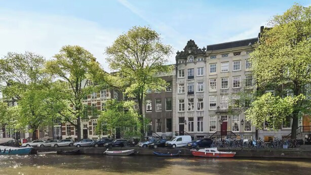 Een gracht met afgemeerde boten, omringd door bomen en historische gebouwen met veel ramen en balkons.