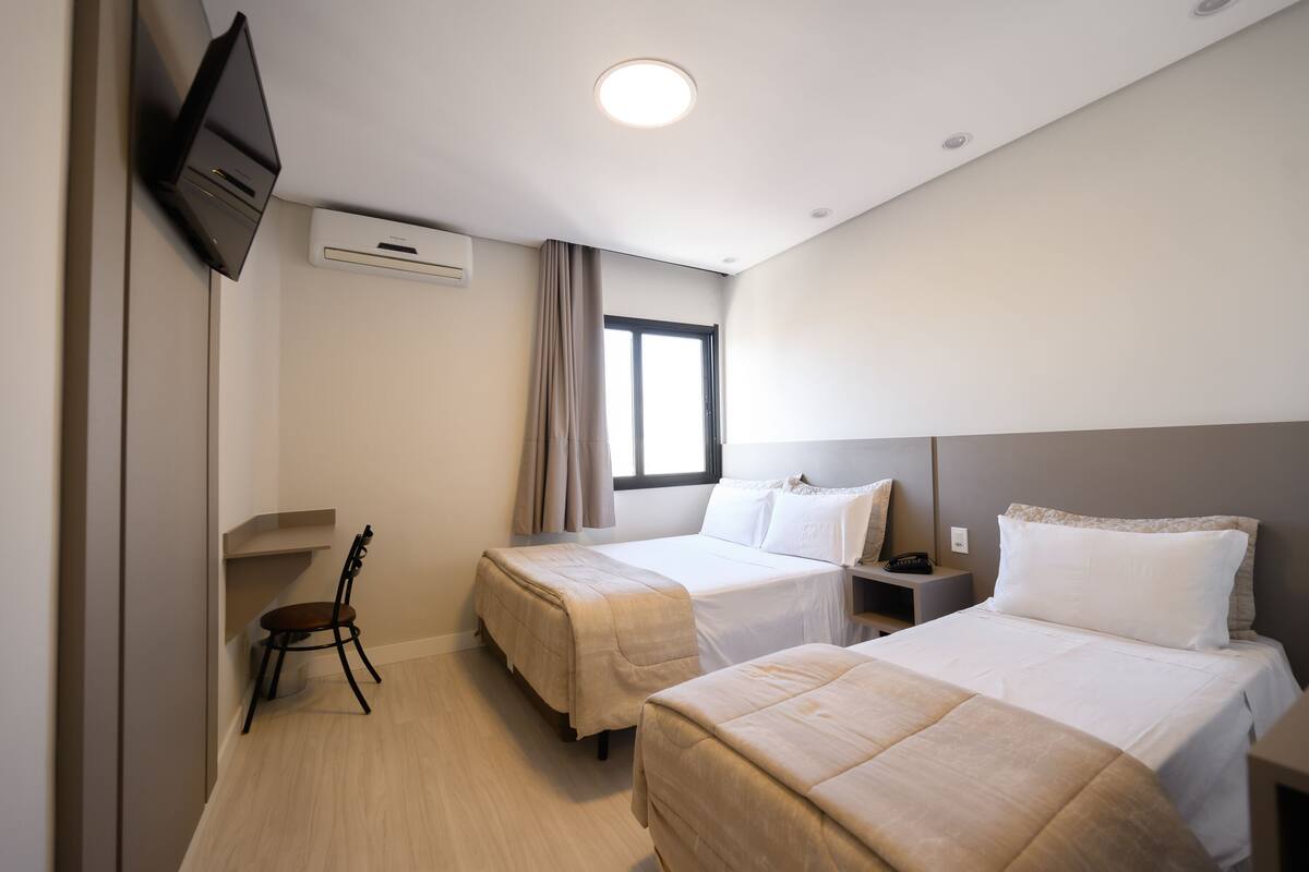 A hotel room with two beds, a TV mounted on the wall, an air conditioning unit, a window with curtains, and a small table with a chair.
