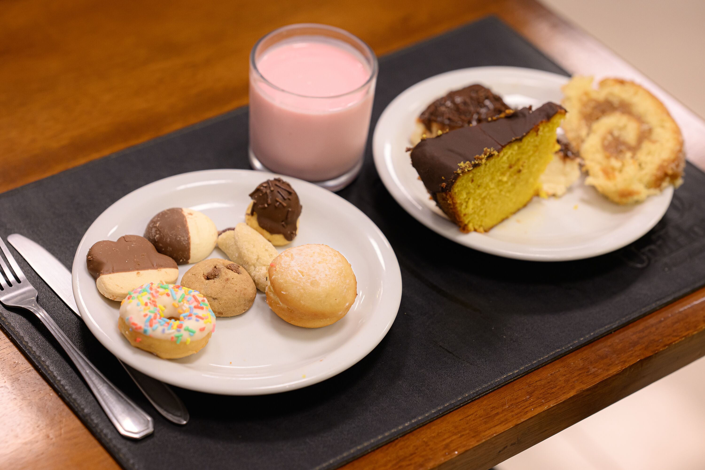 A tray with a plate of assorted cookies and a glass of pink drink, and another plate with a slice of cake and a roll.
