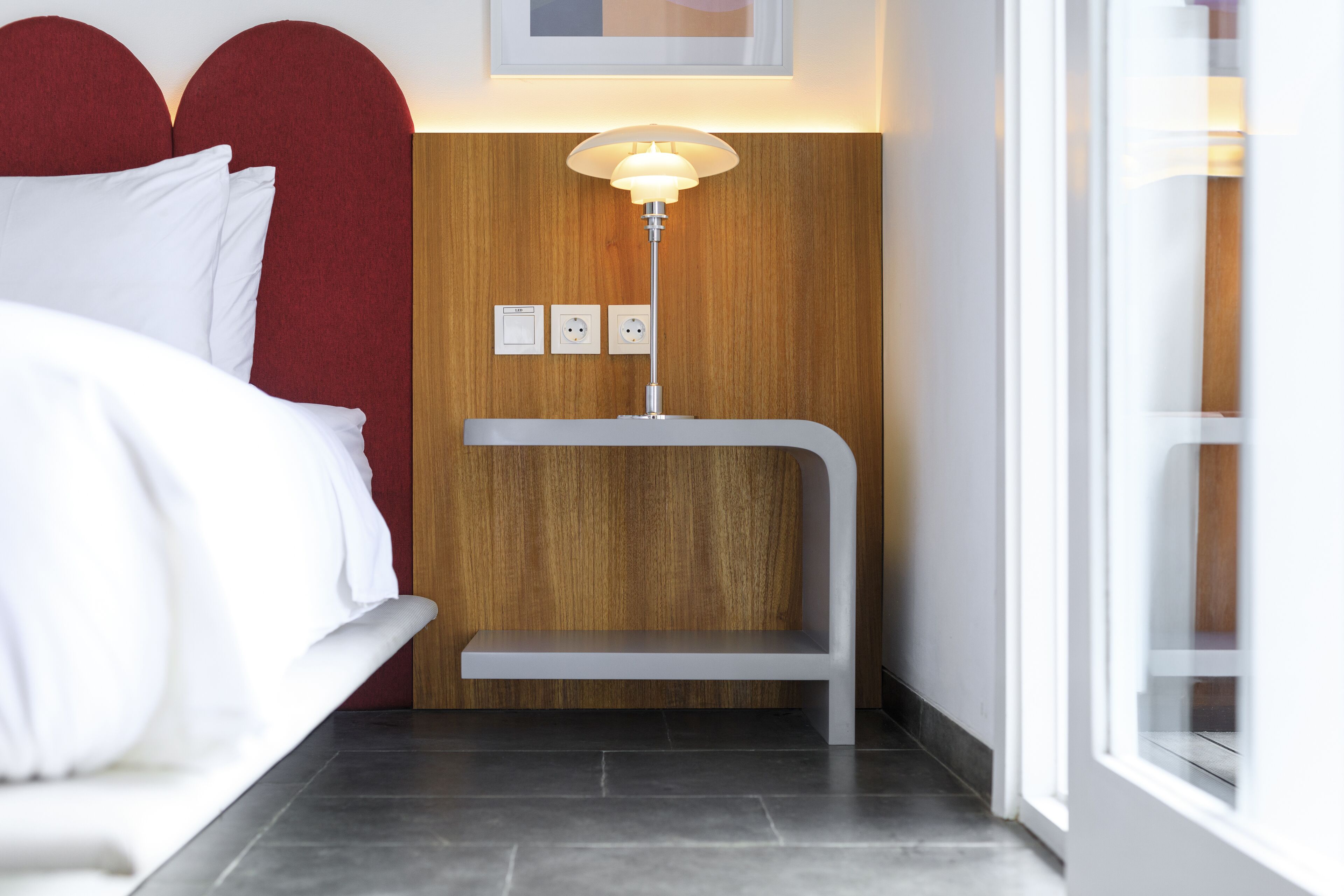 Quarto de hotel moderno com cama, painel de parede em madeira, mesa de cabeceira com luminária e vista para o exterior.