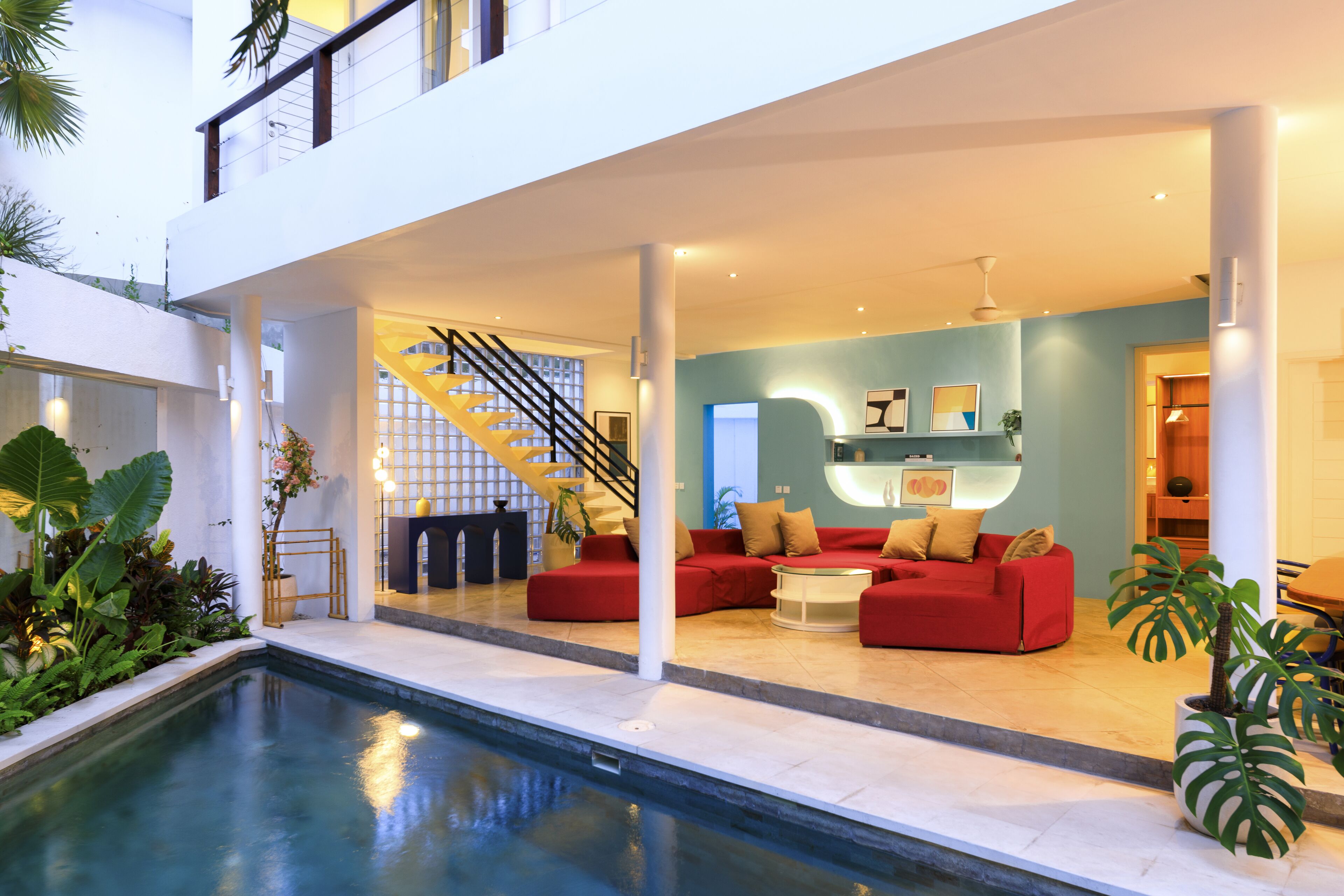 A modern indoor pool area with a red sofa set, a staircase, and a large plant.