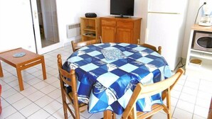 A dining area with a round table covered by a blue and white checkered tablecloth, surrounded by wooden chairs. A television is mounted on a wooden cabinet, and a white refrigerator is visible in the background.