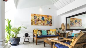 A spacious living room with a sofa, coffee table, and wicker chairs, featuring a large painting and a mirror on the wall. - Villa Atma (Seminyak)