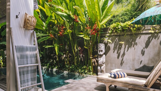 Una terraza de madera con piscina, rodeada de exuberante vegetación tropical y una estatua de piedra.