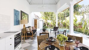 Amplio espacio interior con una mesa de comedor lista para cuatro personas, rodeada de sillas, un sofá y una pequeña mesita auxiliar. Se ve una zona de cocina a la izquierda, y una piscina al fondo.