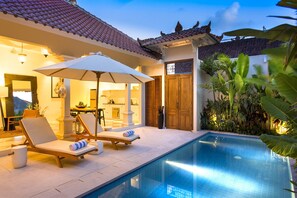 A poolside area with lounge chairs and an umbrella, a kitchen visible through an open door, and a wooden door with a decorative design. - Villa Soleh (Seminyak)