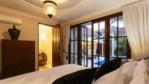 A hotel room with a bed, a wooden wardrobe, a bathroom with a sink, and a view of a pool and outdoor area. - Villa Soleh (Seminyak)