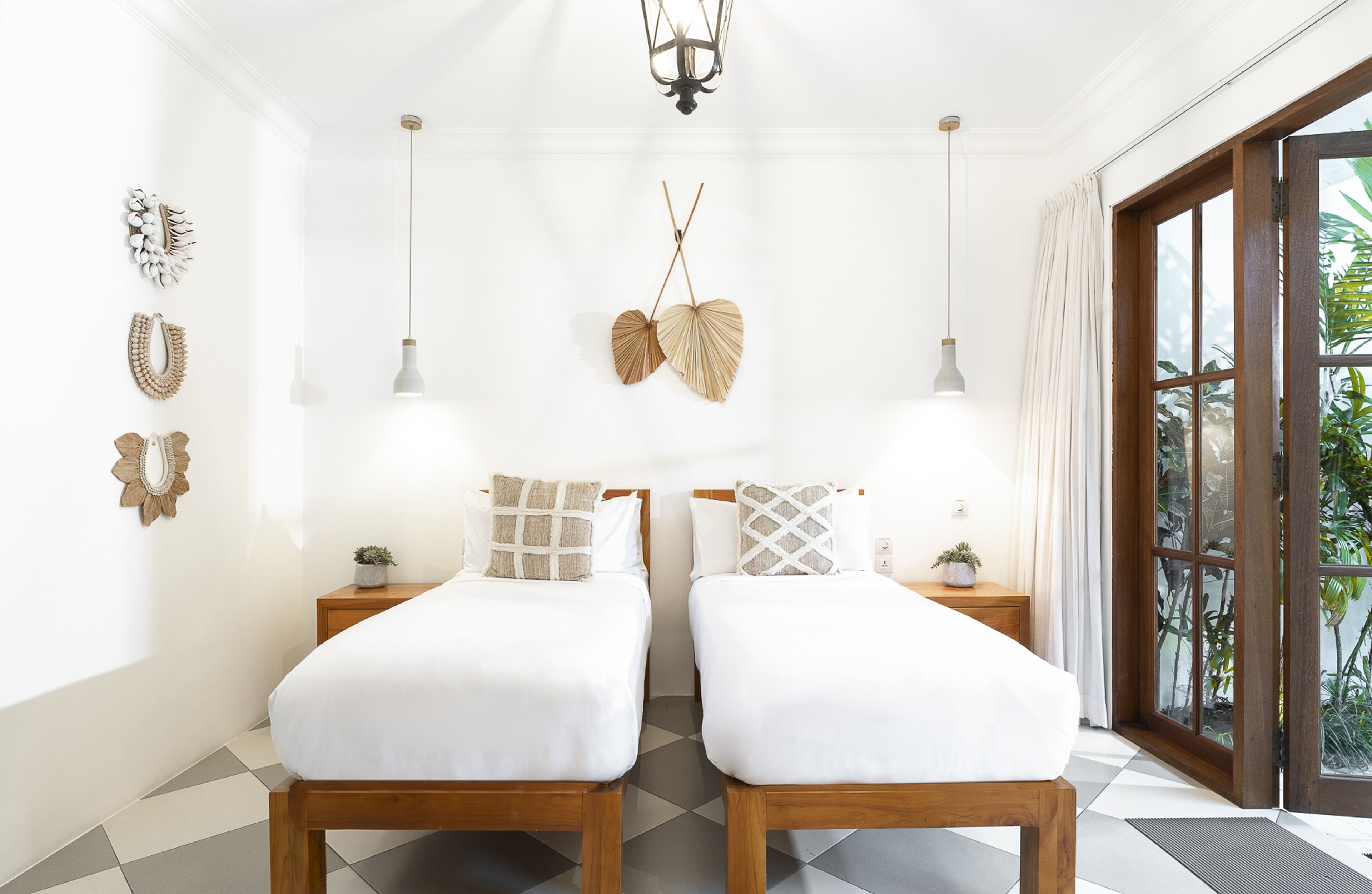 A room with two beds, a chandelier, wall decor, and a view of greenery through a window.