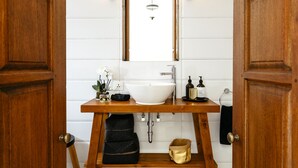 Un baño con tocador de madera, lavabo blanco y un espejo encima. Hay un perchero y un estante pequeño con artículos de aseo.