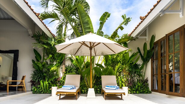 Un área junto a la piscina con dos sillas de descanso, una sombrilla grande y exuberante follaje tropical.