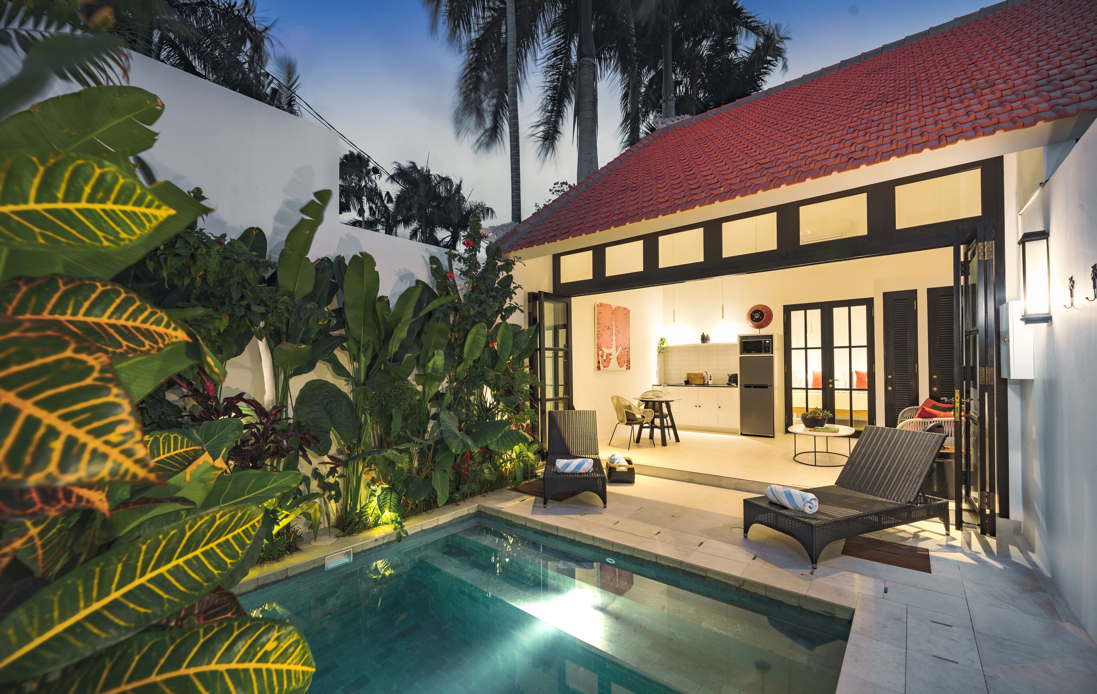 A poolside area with a lounge chair and a dining table, surrounded by tropical plants.