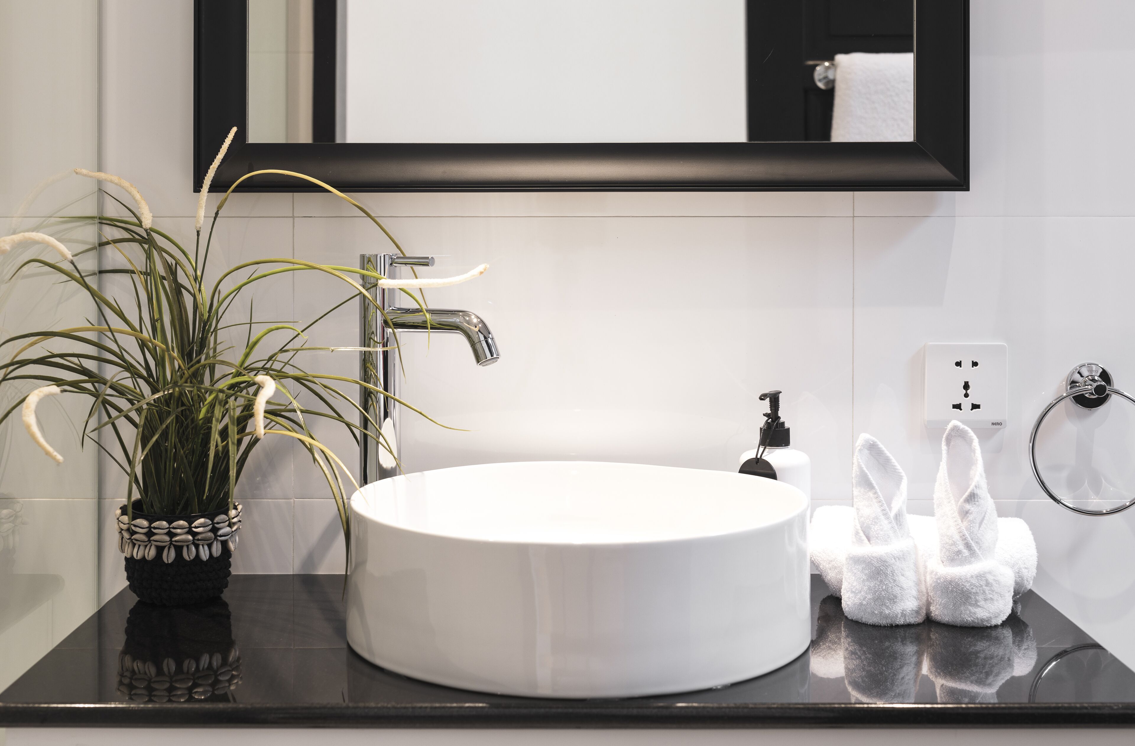 A bathroom with a round white sink, a black countertop, a potted plant, a towel rack, and a mirror above the sink.