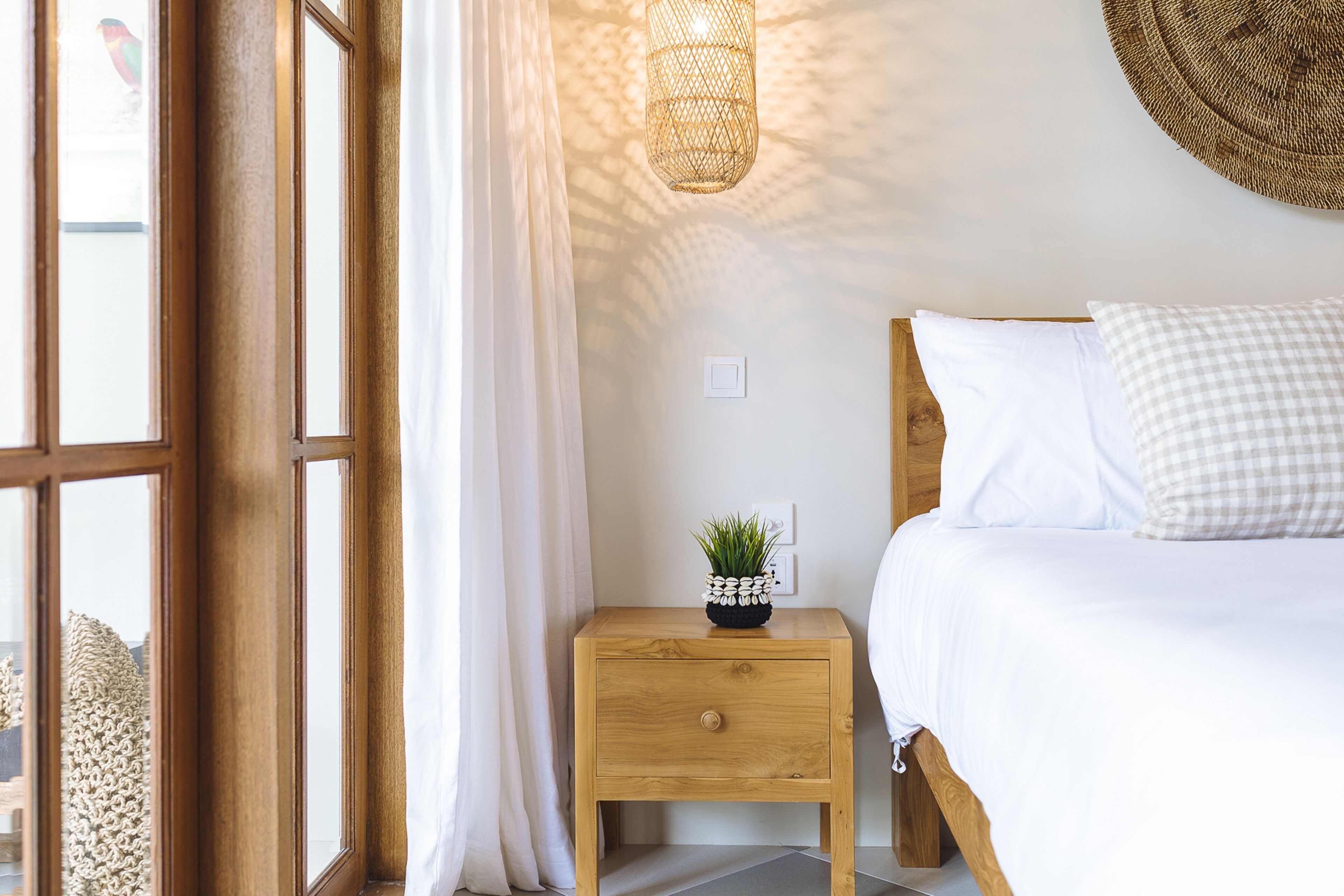 A bedroom with a wooden nightstand, a bed with white bedding, a small potted plant, and a woven light fixture.