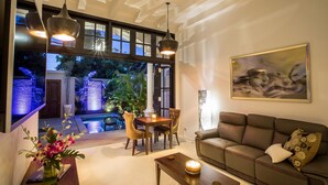 A modern living room with a sofa, a coffee table, and a dining area with a table and chairs. There is a large window with a view of a pool and greenery outside. - Pulau Rumah (Seminyak)