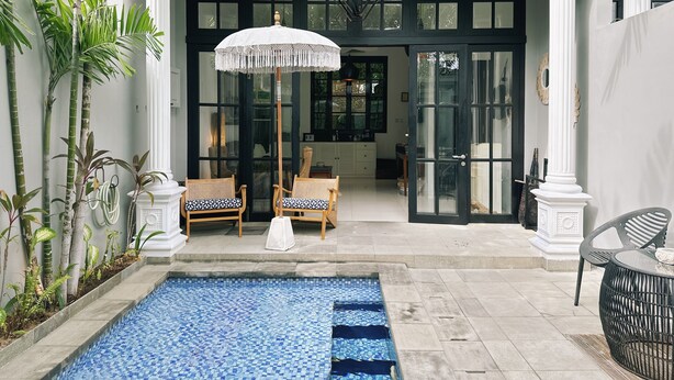 A pool area with a tiled pool, two wicker chairs, a white umbrella, and a view of the interior through glass doors. - Pulau Rumah (Seminyak)