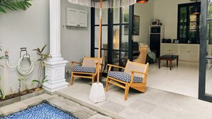 A modern living room with a large glass door leading to a balcony, two wooden chairs with patterned cushions, a white umbrella, and a small pool area. - Pulau Rumah (Seminyak)