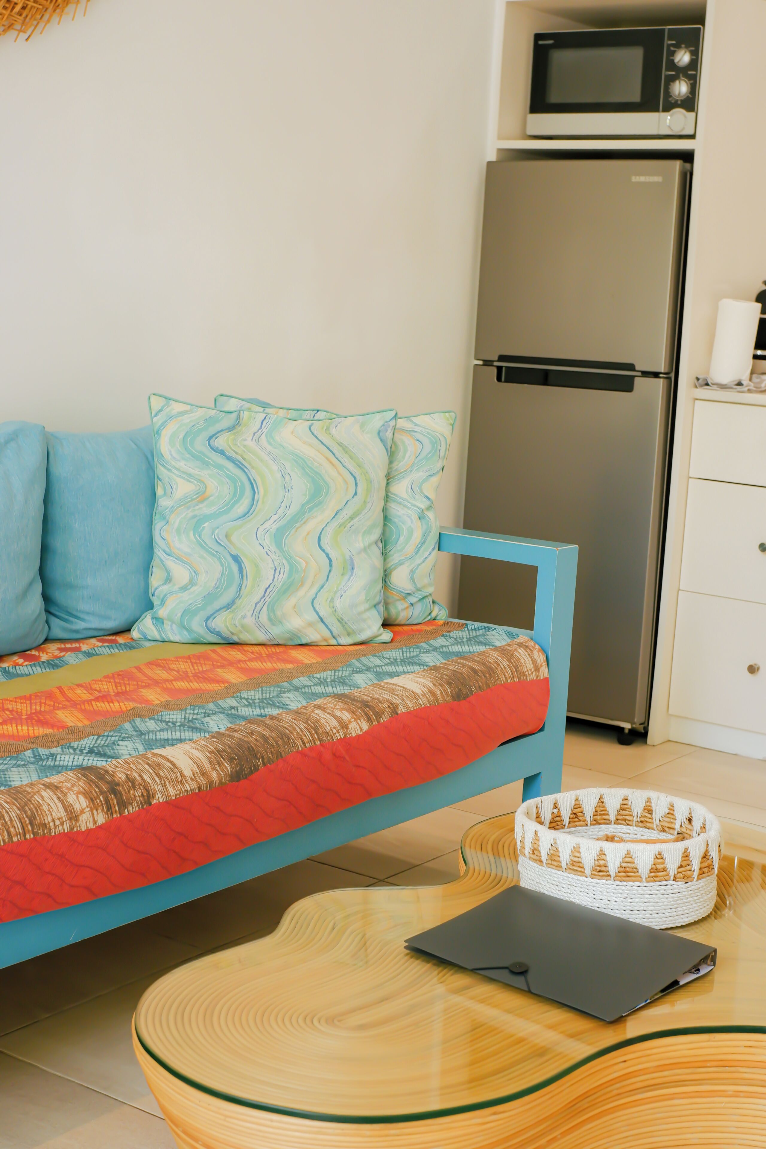 A bed with a blue frame, a patterned quilt, and two pillows. A microwave and refrigerator are visible in the background.