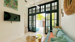 A modern living room with a flat-screen TV, a large window with a view, a ceiling fan, and a woven wall decoration. - Villa Neno (Seminyak)