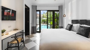 A hotel room with a large bed, a desk with a chair, a TV displaying 'NETFLIX', and a view of a swimming pool and greenery. - Villa Nago (Seminyak)