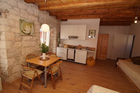 A rustic room with a stone wall, wooden ceiling, a dining table with chairs, a kitchen area with cabinets, and a bed.