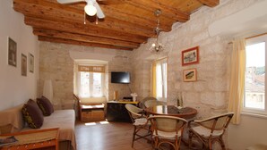 A cozy room with a wooden ceiling, stone walls, a sofa, a dining table with chairs, a TV, and windows with curtains. - Apartments Stelina (Korcula)