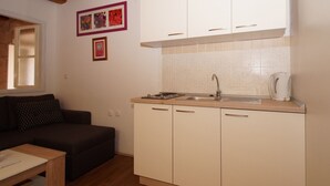 A compact kitchen with white cabinets, a sink, and a coffee maker. There is a dark-colored sofa and a small wooden table in the living area. - Apartments Stelina (Korcula)