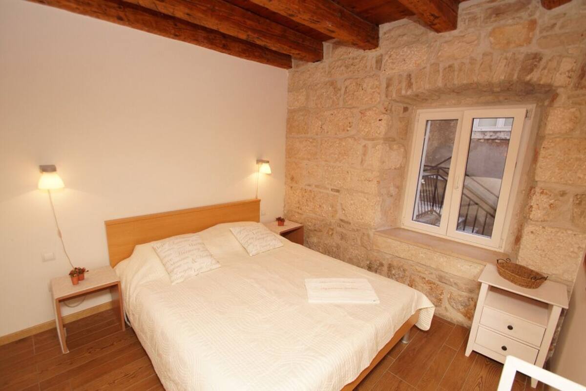 A bedroom with a wooden bed, stone walls, a window with a balcony, and a small bedside table with a lamp.