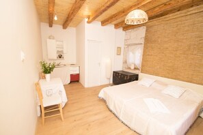 A bedroom with a wooden ceiling, a bed with white bedding, a bedside table with a plant, and a small desk with a chair.