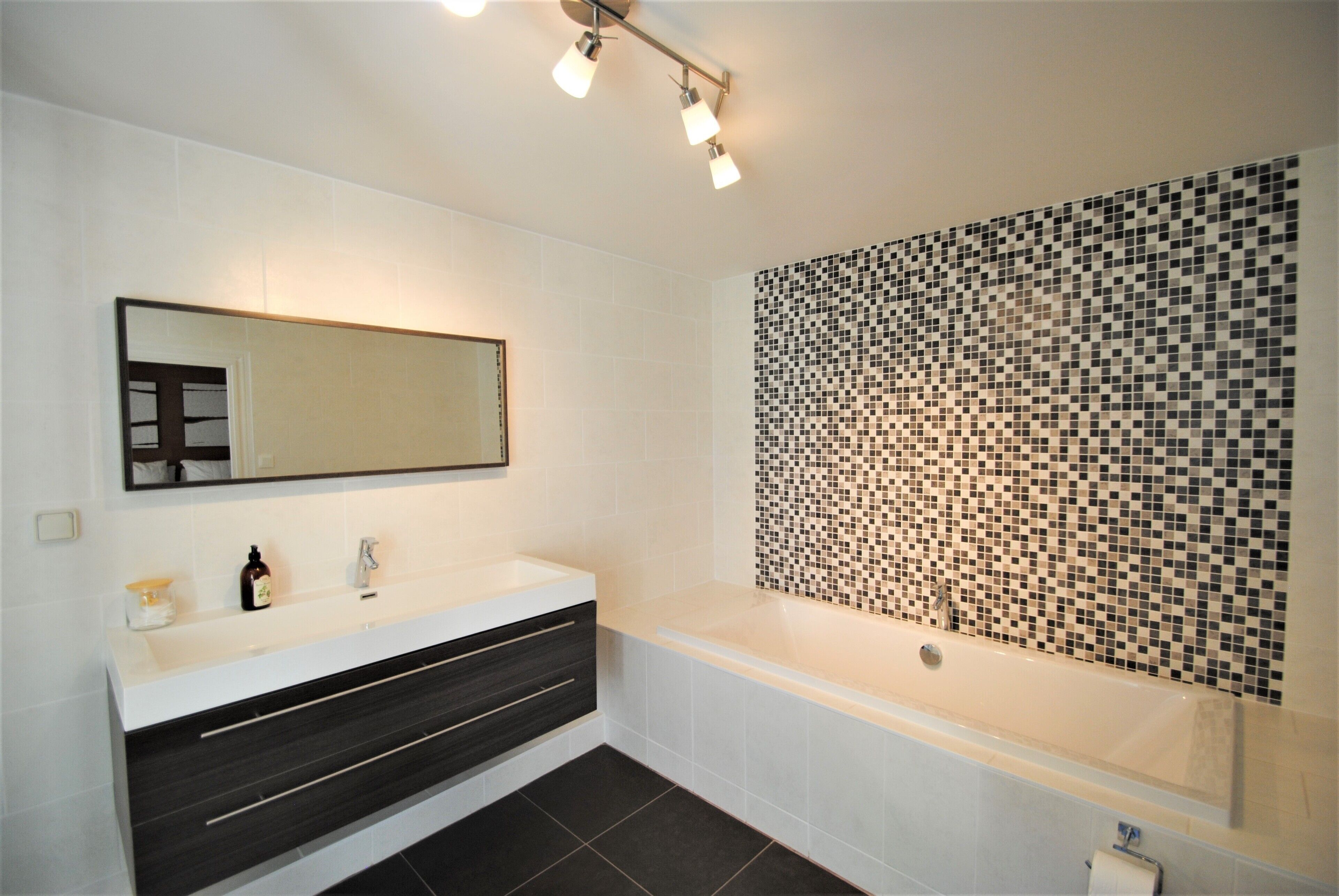 A modern bathroom with a white vanity, dark drawers, a large mirror, and a mosaic tile wall.