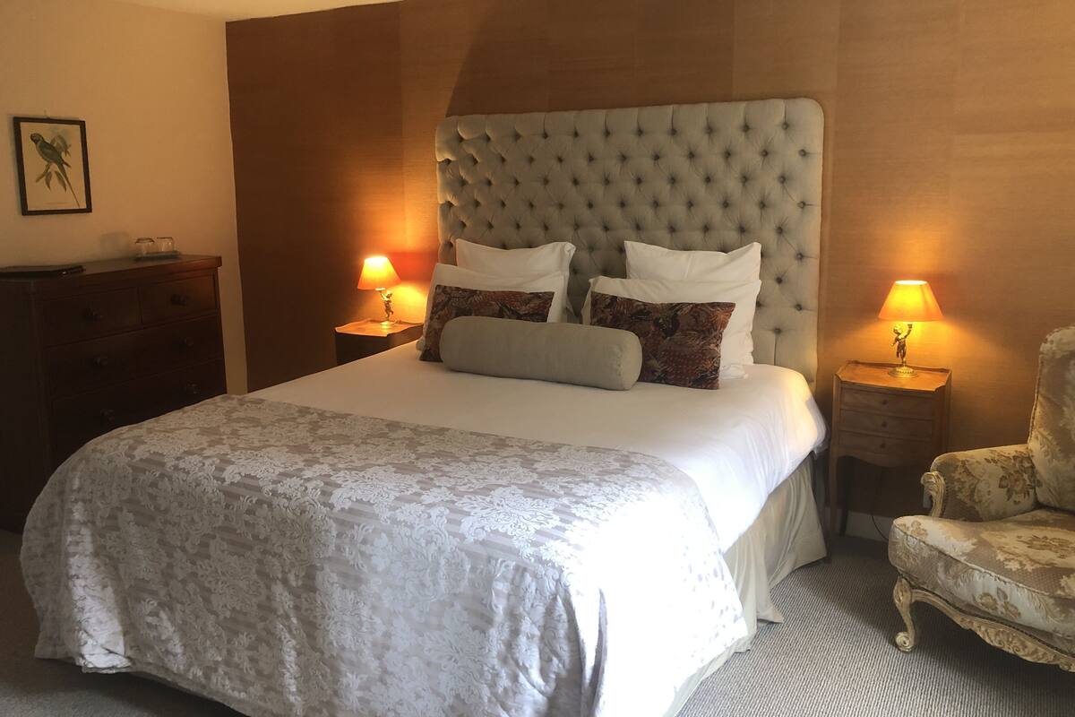 Een net opgemaakt bed met een gestoffeerd hoofdbord, twee bedlampen, een houten dressoir en een fauteuil met bloemenprint.
