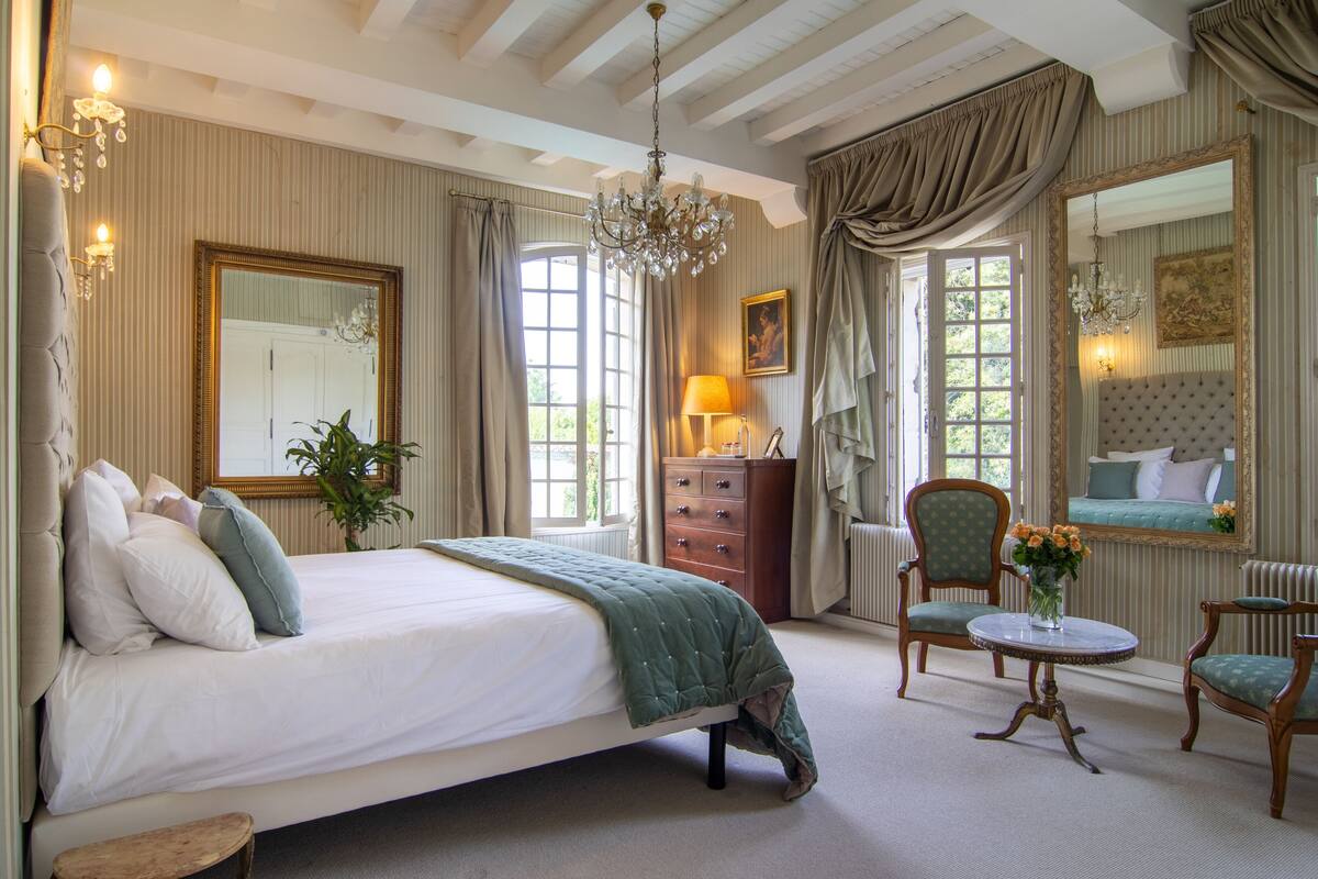 Een ruime slaapkamer met een groot bed, een kroonluchter, een kaptafel en een zithoek met stoelen en een tafeltje.