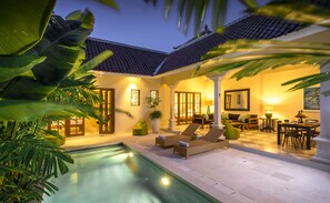 A well-lit outdoor area with a pool, lounge chairs, and a dining table set for two. - Villa Mandala (Seminyak)