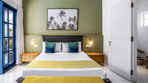 A double bed with a yellow bedspread, two nightstands with wall-mounted lamps, a framed picture of palm trees, and a blue-framed window. - Beachhouse (Canggu)