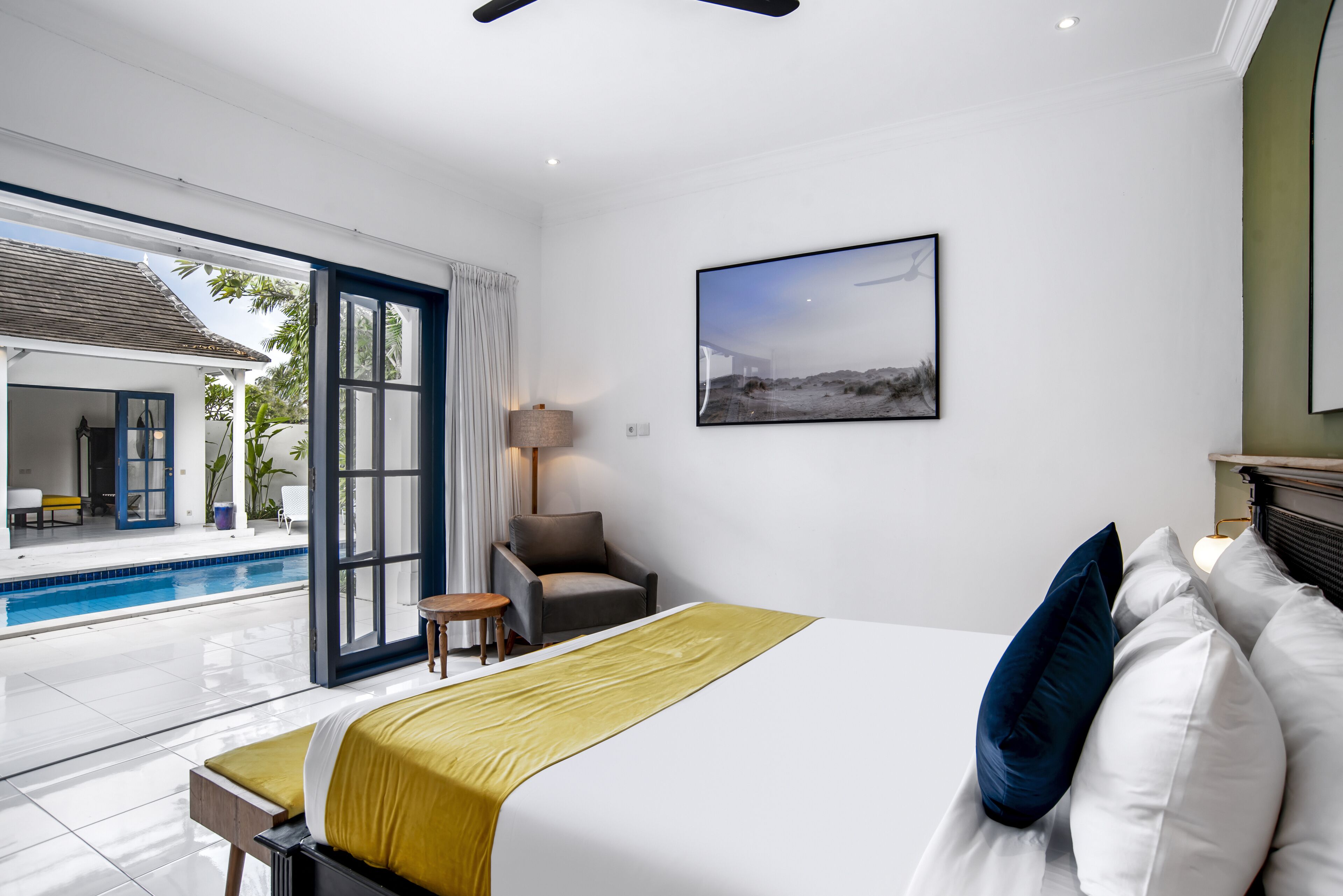 A hotel room with a bed, a chair, a small table, a lamp, and a view of an outdoor swimming pool.