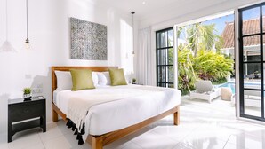 A spacious bedroom with a large bed, a nightstand, a wall art piece, and a view of a pool and greenery outside. - Town House (Canggu)