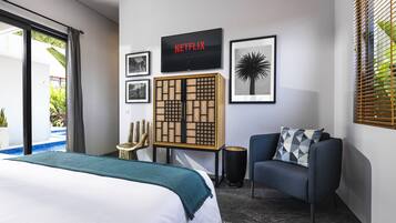 A modern hotel room with a bed, a blue armchair, a wall-mounted TV displaying 'NETFLIX', and framed pictures.