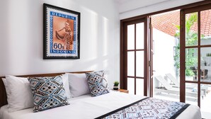 A neatly made bed with patterned pillows, a framed Indonesian stamp on the wall, and a view of a balcony with outdoor furniture. - Villa Salsa (Seminyak)