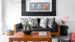 A living room with a black-framed picture above a black sofa, a wooden coffee table with a pineapple bowl, and a vase with pink flowers. - Villa Salsa (Seminyak)