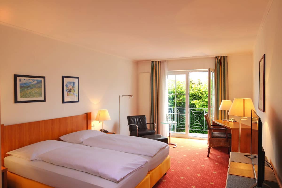 Ein Hotelzimmer mit einem großen Bett, einem Schreibtisch, einem Sessel und einem Balkon mit Blick auf Grünflächen.
