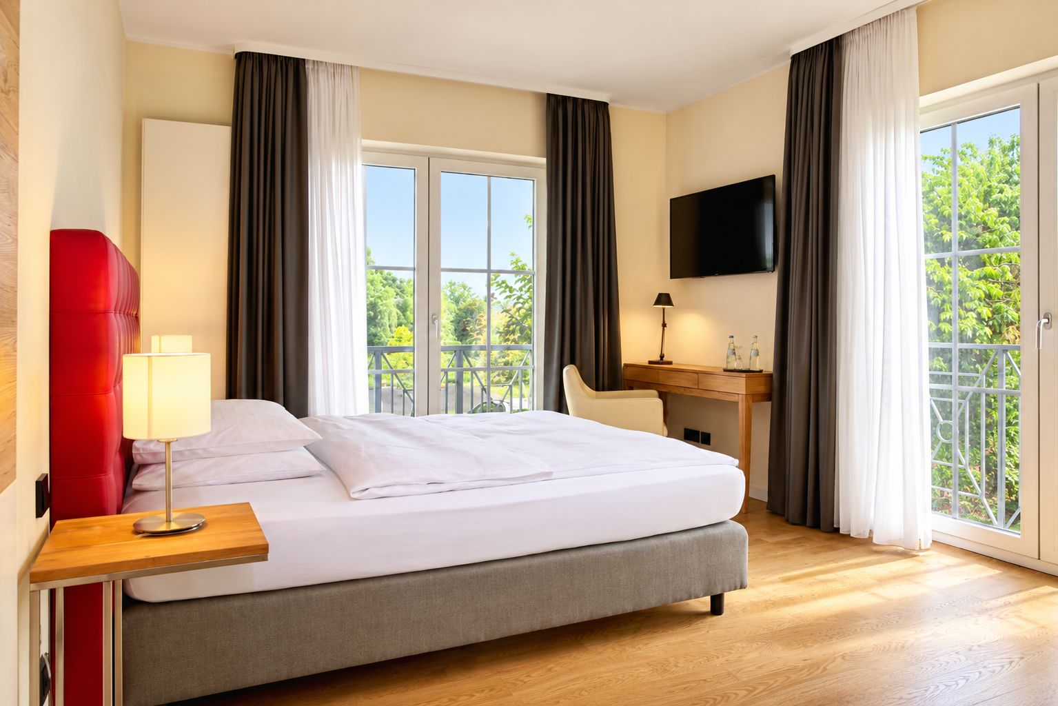A hotel room with a bed, a desk, a TV, and a balcony with a view of trees.