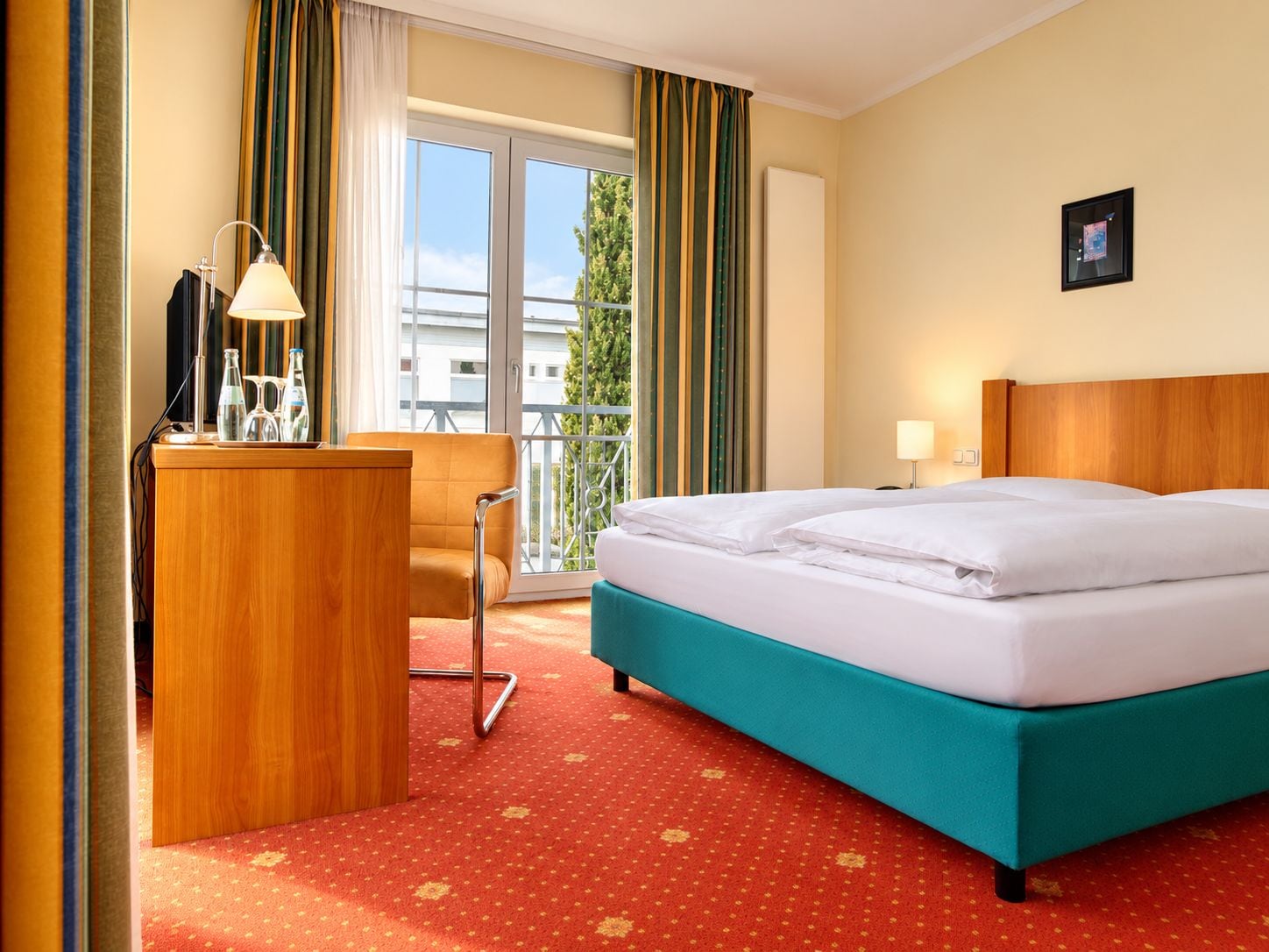 A hotel room with a bed, a desk, a chair, a window with curtains, and a view of buildings.