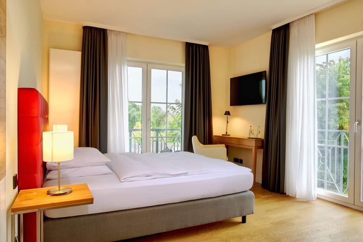 Ein Hotelzimmer mit Bett, Schreibtisch, Fernseher und einem Balkon mit Vorhängen.