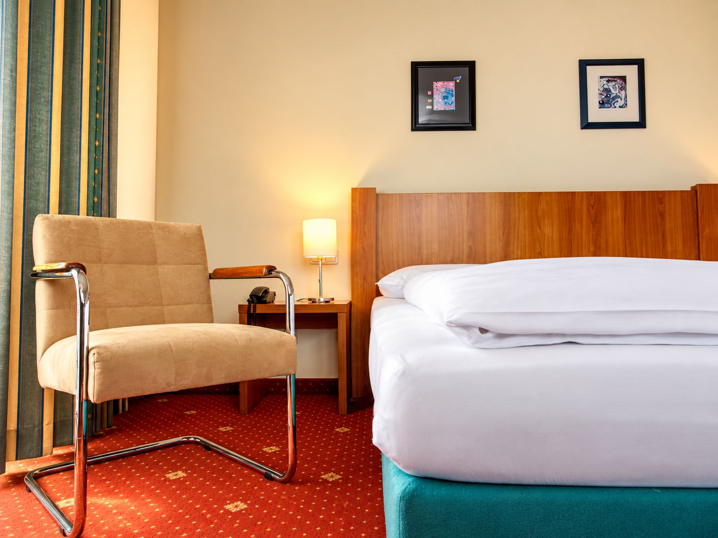 A hotel room with a bed, a chair, a nightstand, a lamp, and two framed pictures on the wall.