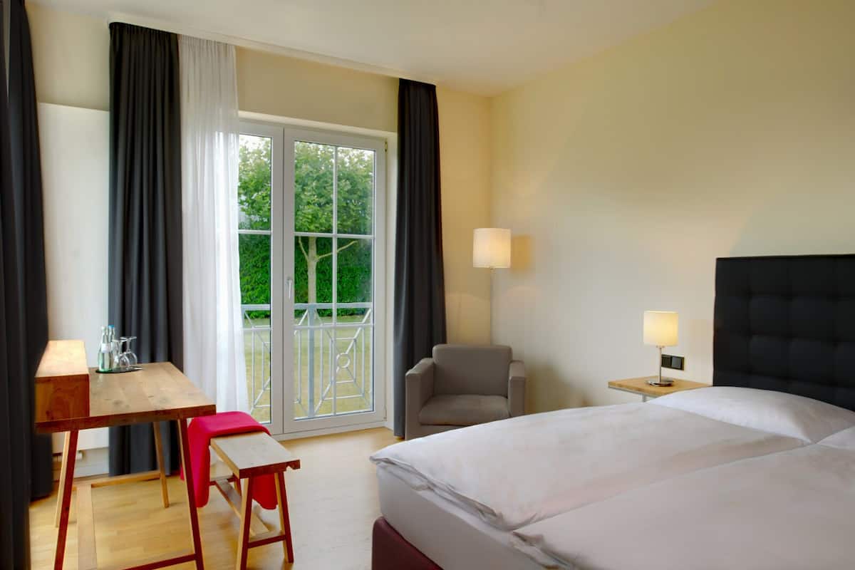 Ein Hotelzimmer mit Bett, Schreibtisch, Stuhl, Lampe und Blick auf Grün hinter einer Schiebetür.