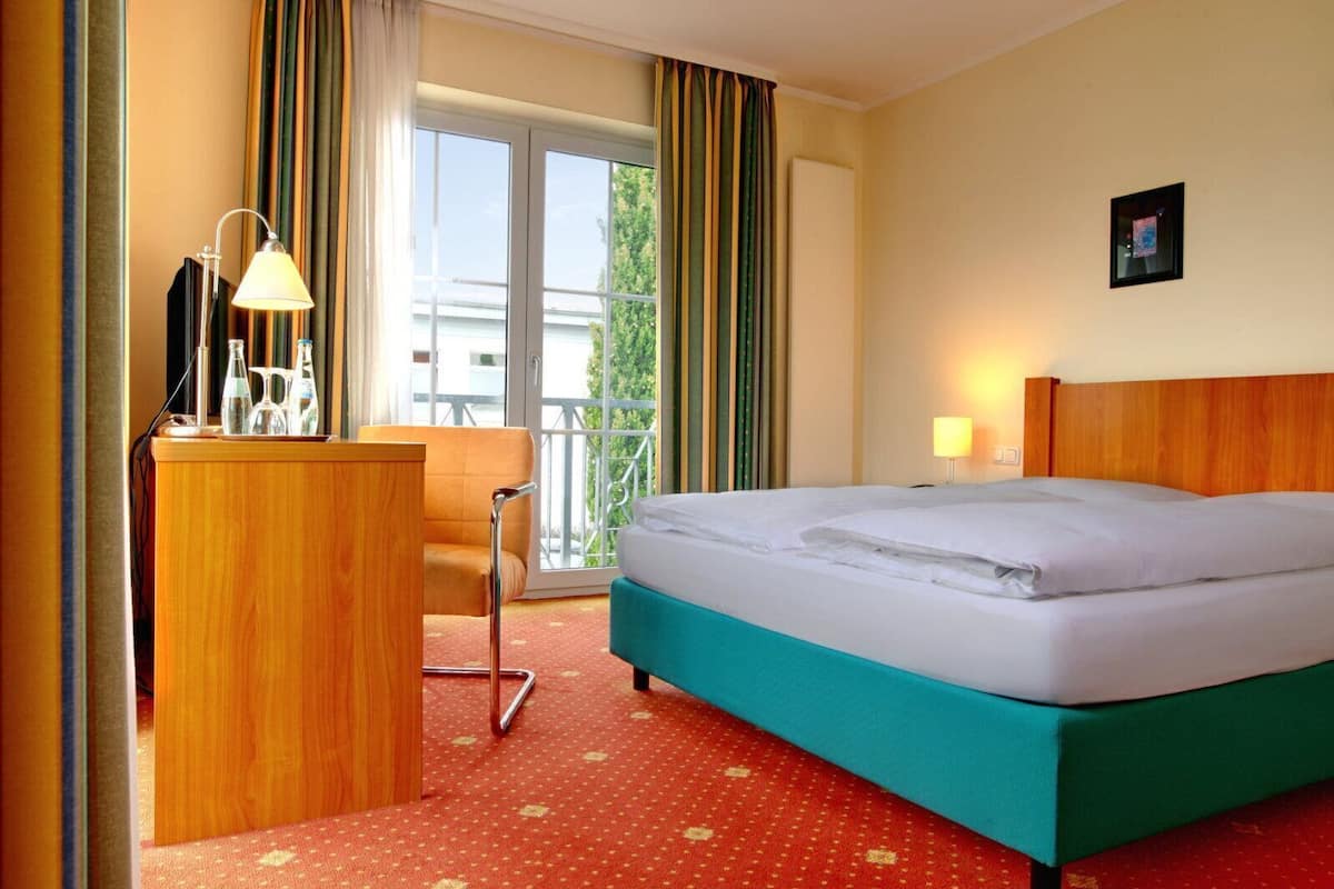 Ein Hotelzimmer mit Bett, Schreibtisch mit Lampe, Stuhl, Vorhängen am Fenster und Blick auf Gebäude.