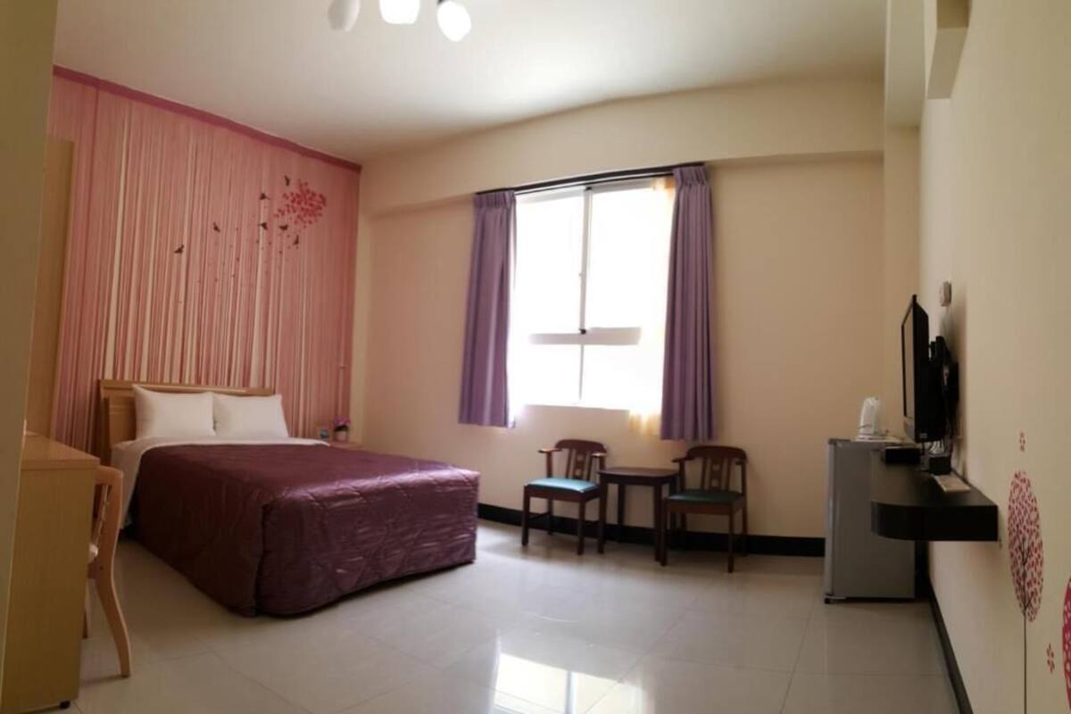 Habitación de hotel con cama, escritorio, silla, mesita, televisor y ventana con cortinas.