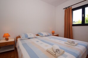Uma cama bem arrumada com colcha xadrez, dois travesseiros, uma mesa de cabeceira com abajur e uma janela com cortinas.